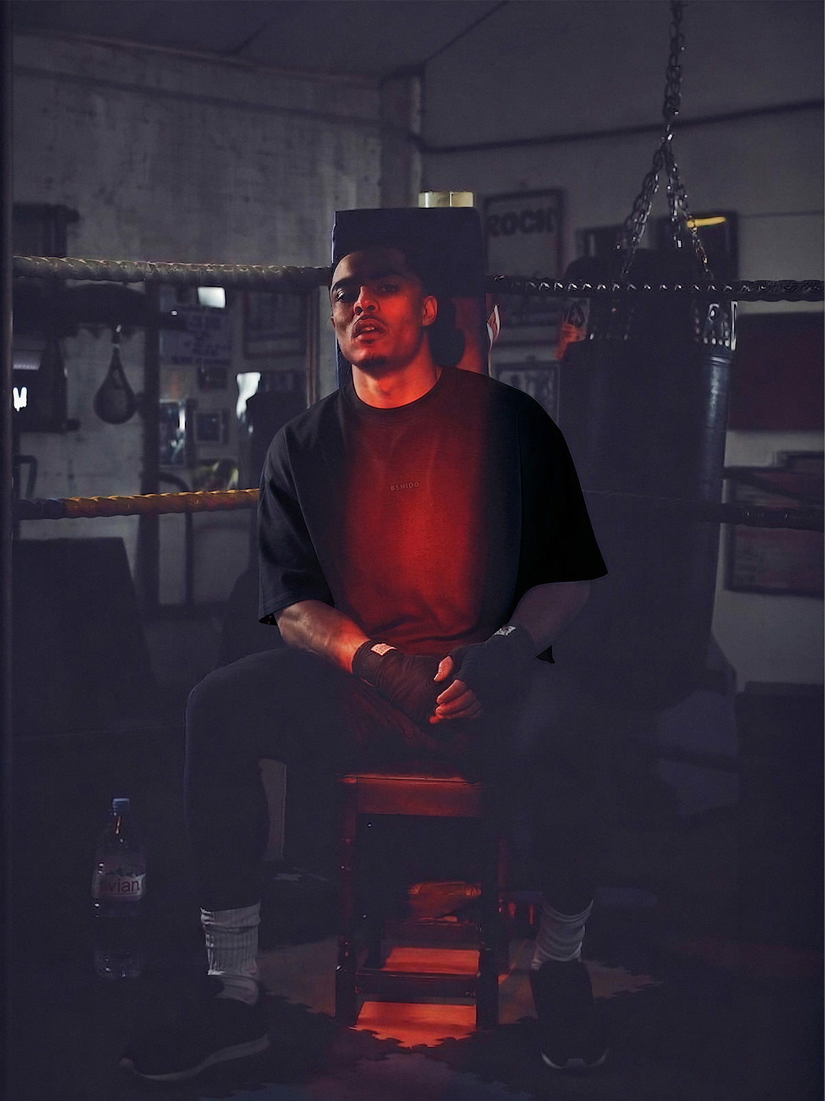 Man wearing Bshidō streetwear sitting in a boxing ring corner with a heavy bag in the background.