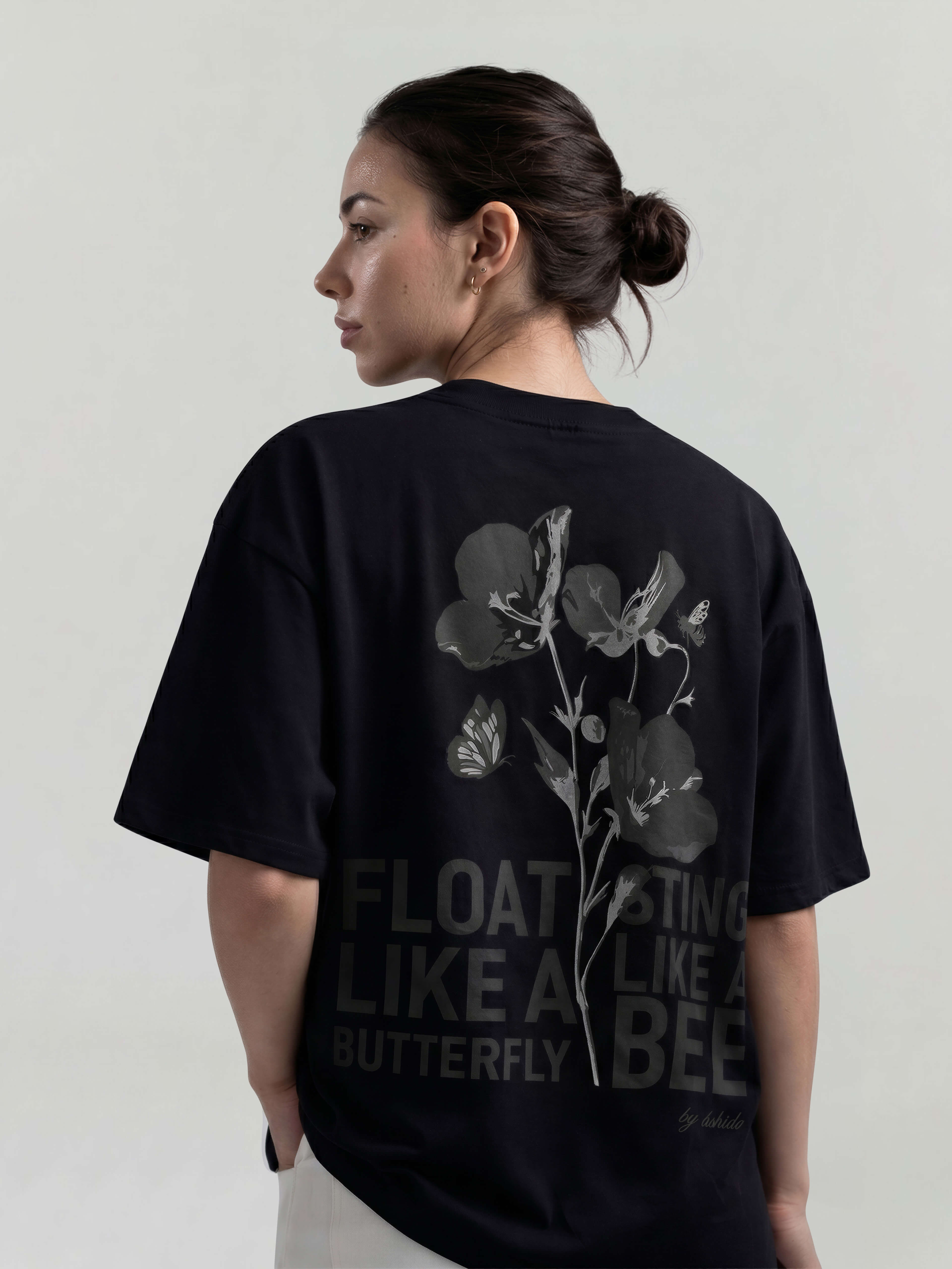 Female model wearing black oversized Bshido t-shirt back angled view with floral graphic and “Float like a butterfly sting like a bee” print