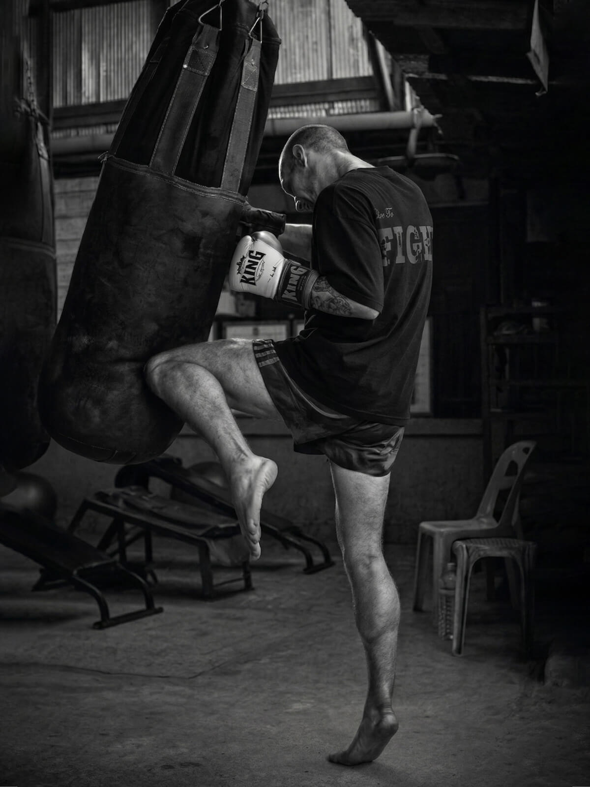 Muay Thai fighter kicking a heavy bag during training in a gym wearing a t-shirt with “Live to Fight” print
