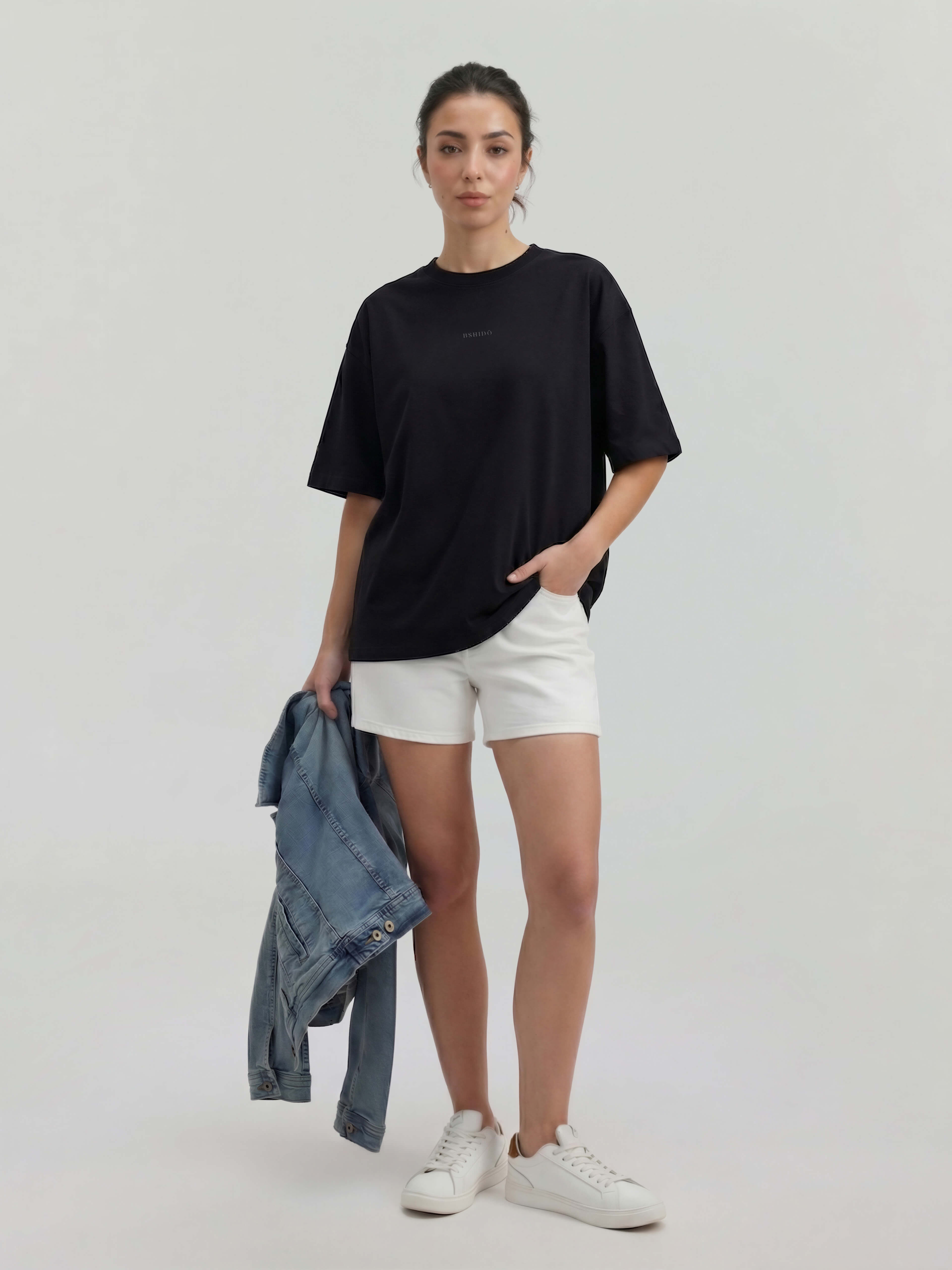 Female model wearing black oversized Bshido t-shirt front view styled with white shorts and holding denim jacket