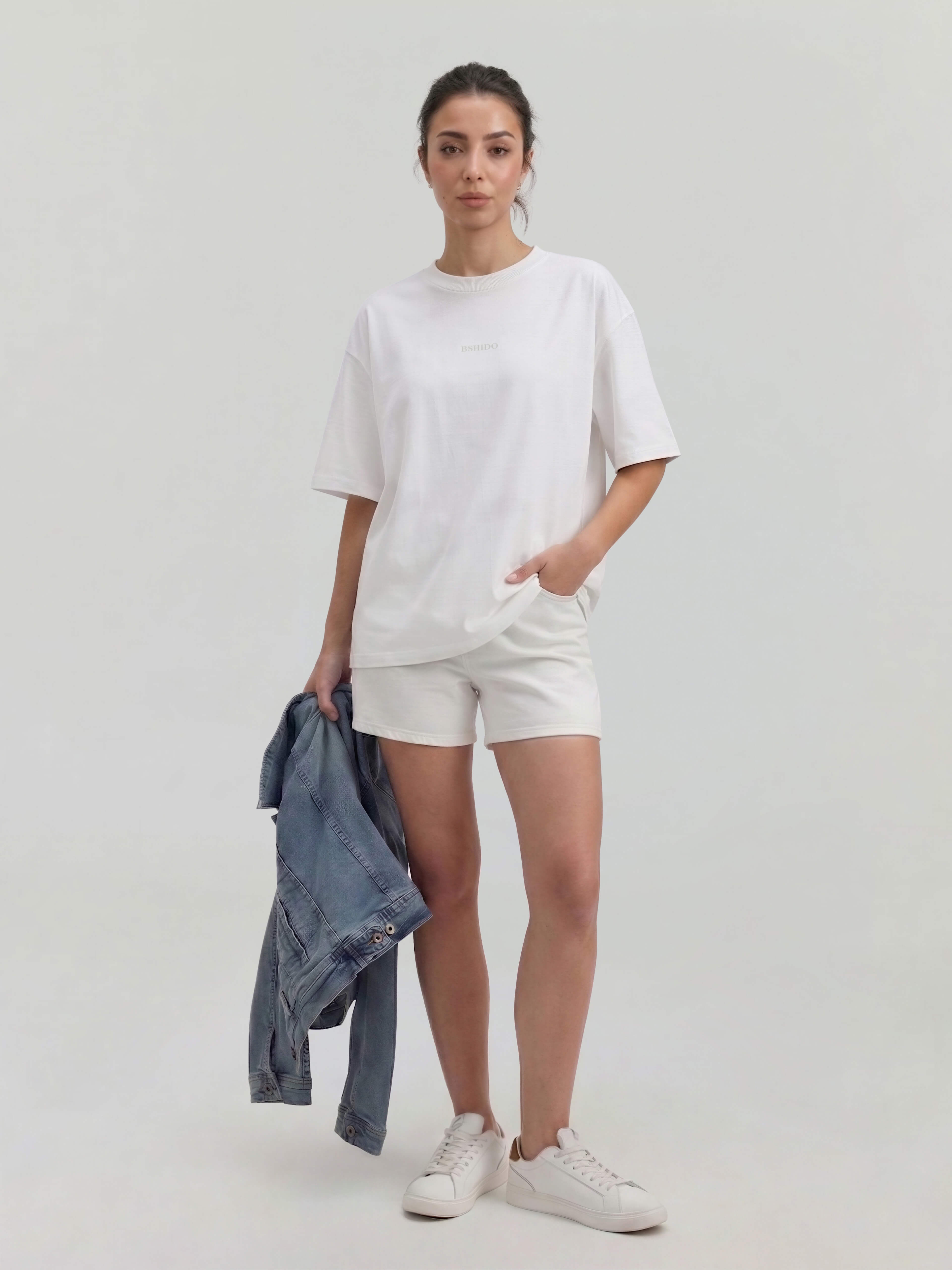 Female model wearing white oversized Bshido t-shirt front full body view styled with white shorts and holding denim jacket
