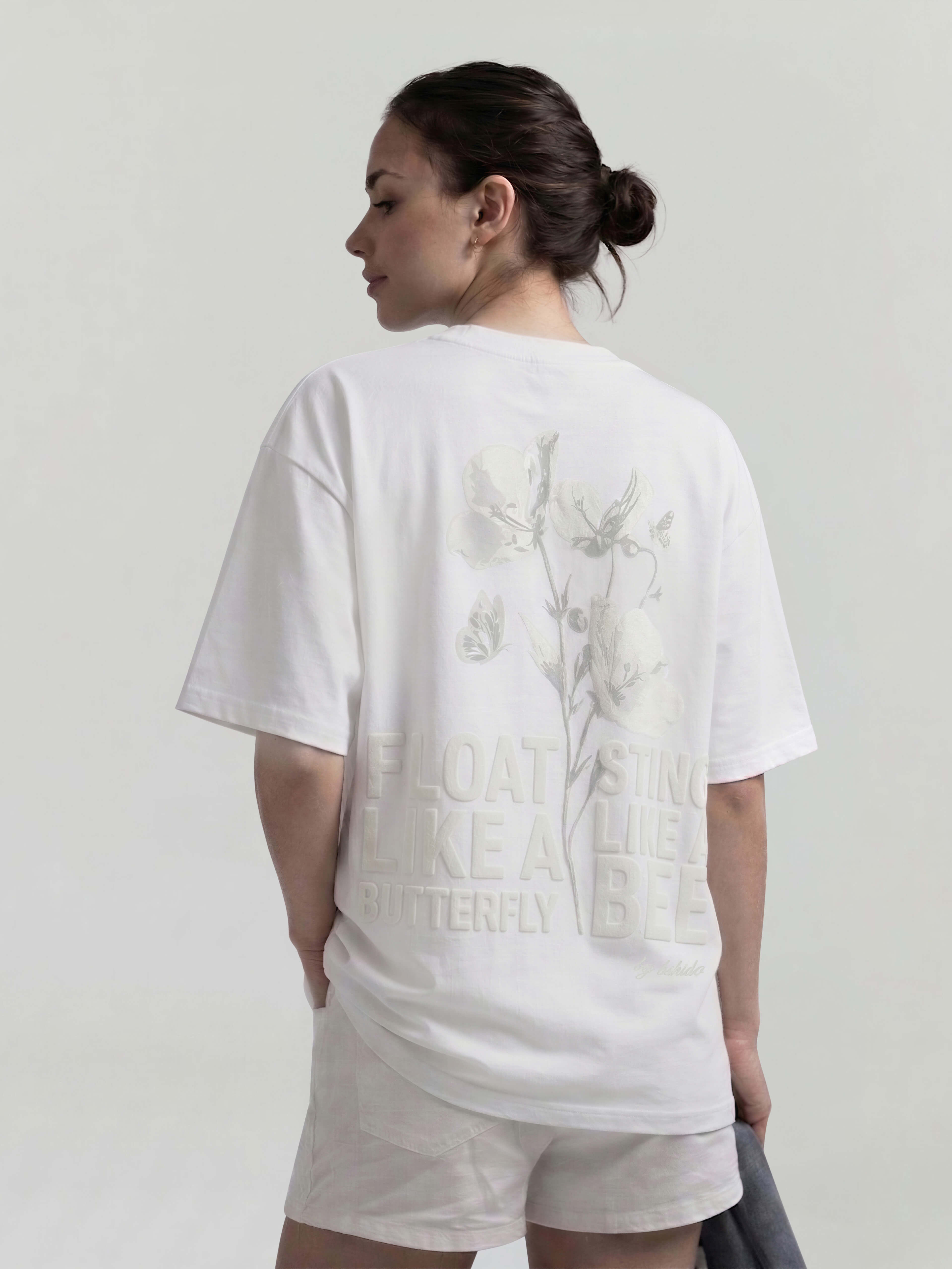 Female model wearing white oversized Bshido t-shirt back angled view with floral graphic and “Float like a butterfly sting like a bee” print