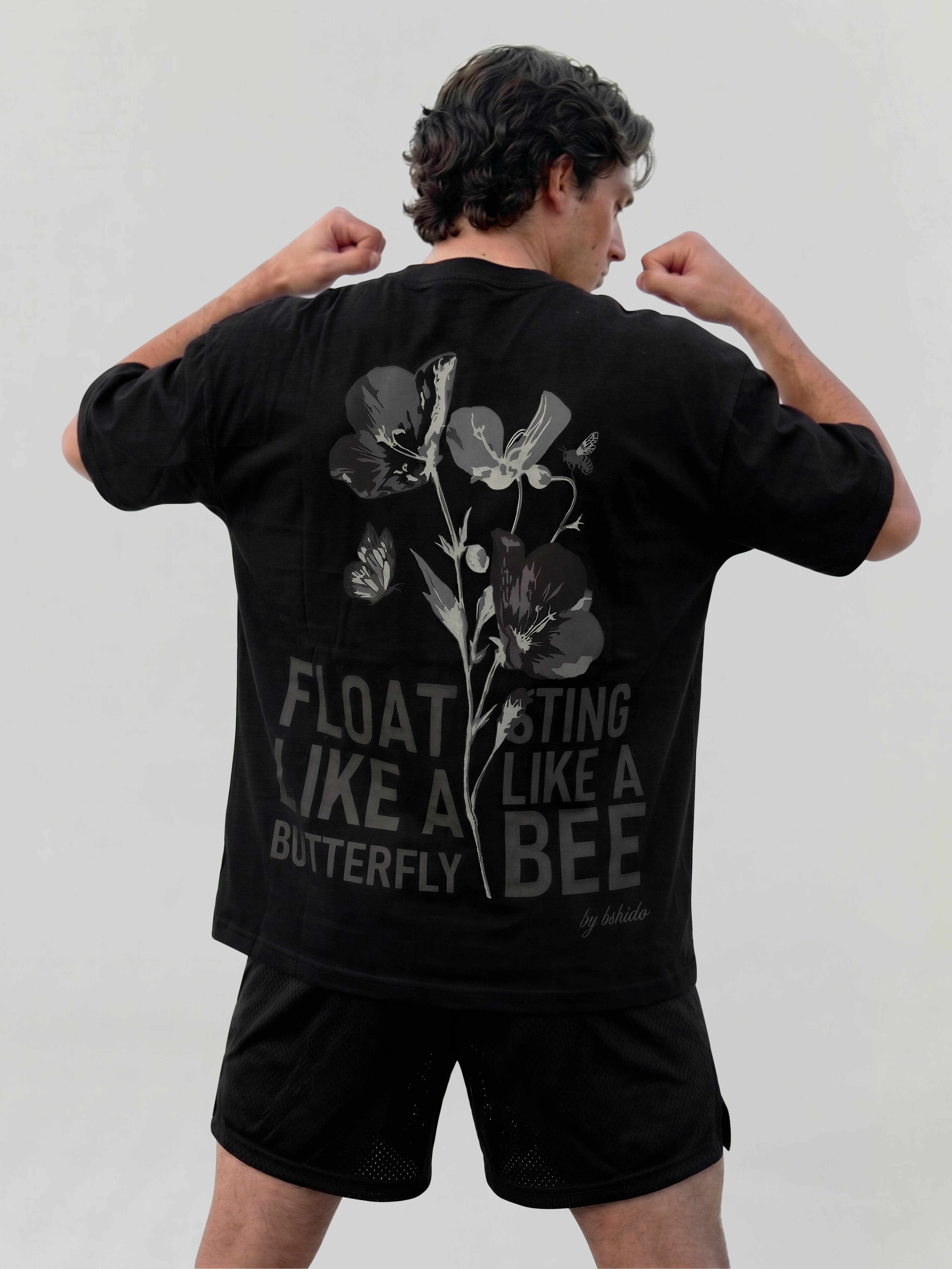 Male model flexing in black oversized Bshido t-shirt showing back graphic with floral design and “Float like a butterfly sting like a bee” text
